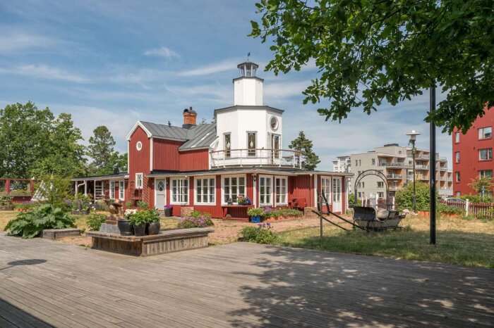 Rött hus med vita knutar, lanternin, veranda, träbrygga, blommor, bänk, urban bakgrund, blå himmel, gröna träd.