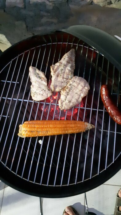 Kolgrill med kycklingfiléer, korv och majs. Utomhus, ser ut som kvällstid. En person i bakgrunden.