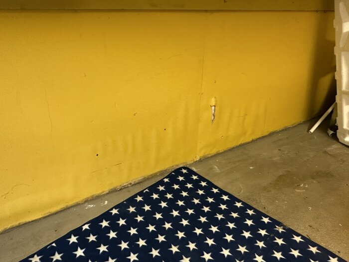 Gul vägg med skador, amerikansk flaggmönstrad matta i förgrunden, inomhusmiljö med betonggolv.