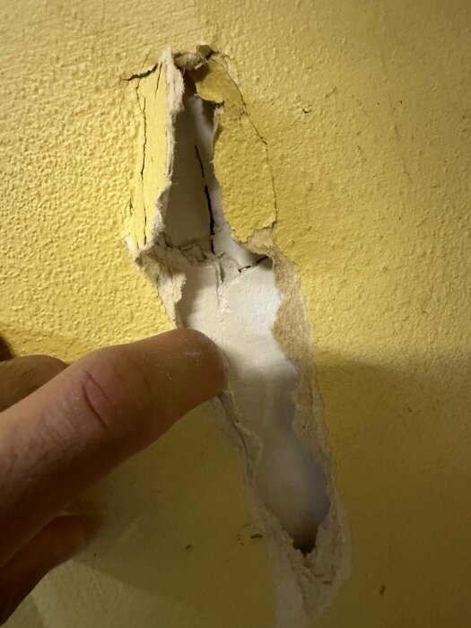 En person pekar på ett hål i en gul vägg, vilket visar skada på ytan.