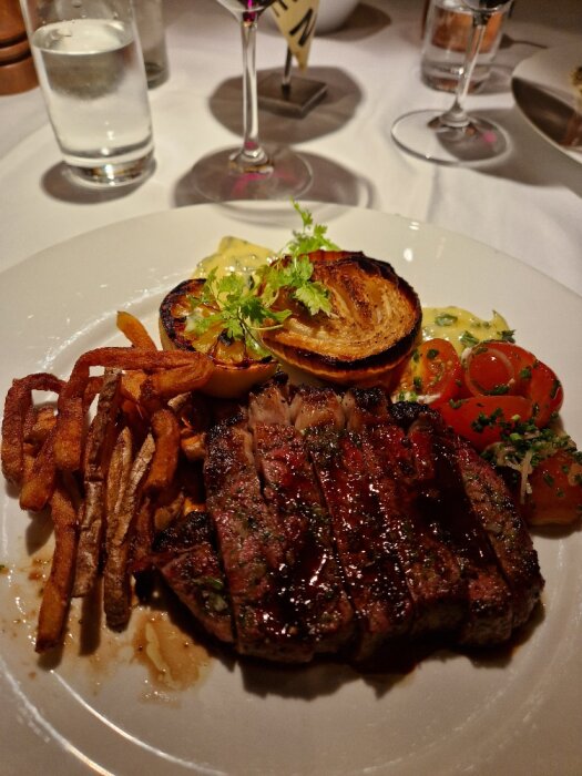 Grillad biff med pommes frites, grillad tomat, lök, bearnaisesås, på en vit tallrik, elegant restaurangmiljö.