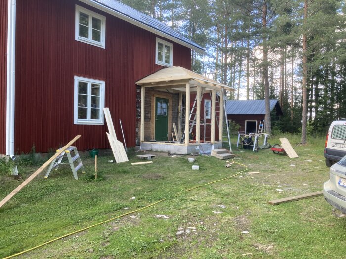 Röd stuga under renovering, material och verktyg utspridda, ny veranda byggs, skogsbakgrund, klar dag.