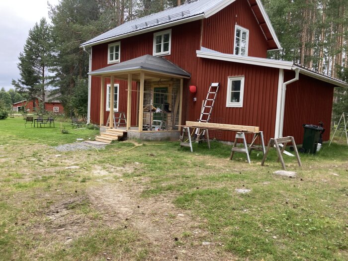 Rött landsbygdshus med vit knut, veranda under arbete, grön gräsmatta, skog bakom, sommar, molnigt, utomhusmöbler.