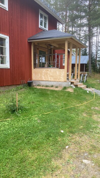 Rött hus med veranda under konstruktion, grönt gräs, träd, stege, sladdar på marken.