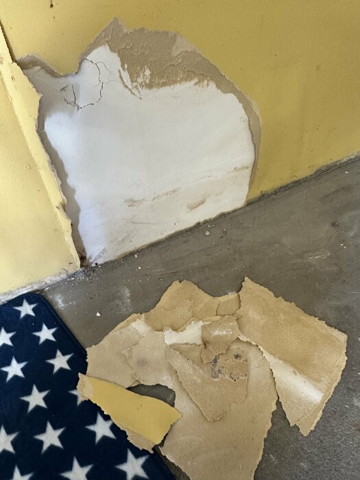 Skadad gipsvägg, gul färg, avfall på golvet, stjärnmönstrad textil. Renoveringsbehov synligt.