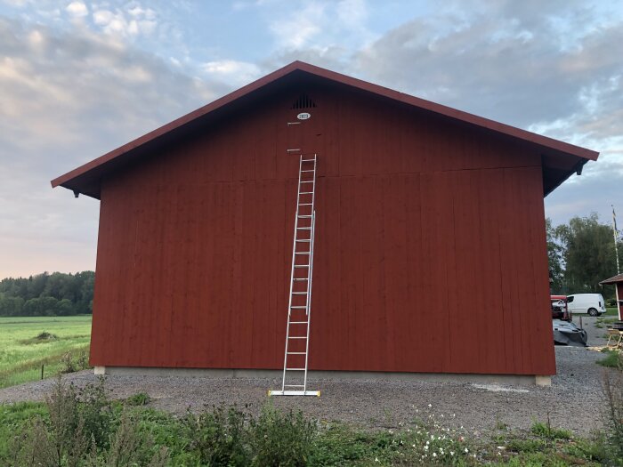 Stor röd lada med stege, grönt fält i bakgrunden, molnig himmel, grus framför.