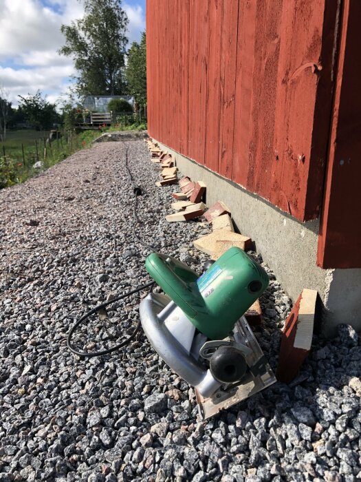 Elektrisk kapsåg på grus vid röd trävägg, murade tegelstenar, trädgård i bakgrunden, soligt, utomhusarbete.