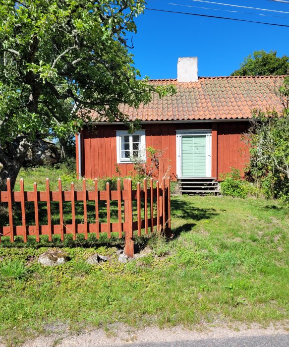 Ett rött trähus med gröna fönsterluckor, tegeltak och ett staket, omgivet av grönska under en klarblå himmel.