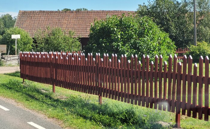 Soligt landskap med rött staket, gröna träd, tegeltak, väg, och en skylt.