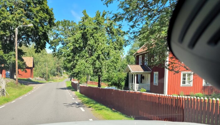 Bild tagen från bil: solig dag, landsväg, gröna träd, röda trähus, typisk svensk bymiljö.