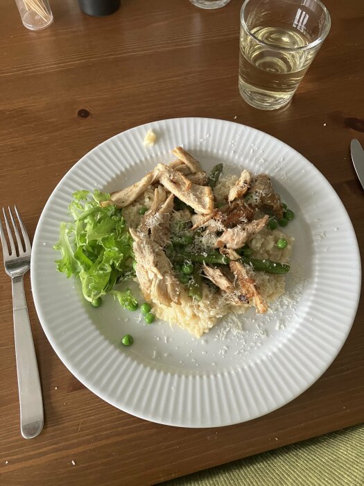 En tallrik med pasta, kyckling, gröna ärtor och sallad, servett, bestick, ett glas vatten på matbord.