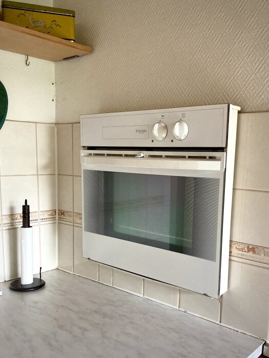 Inbyggd vit ugn i ett kök med kakelvägg, kökshylla och hushållspappershållare.