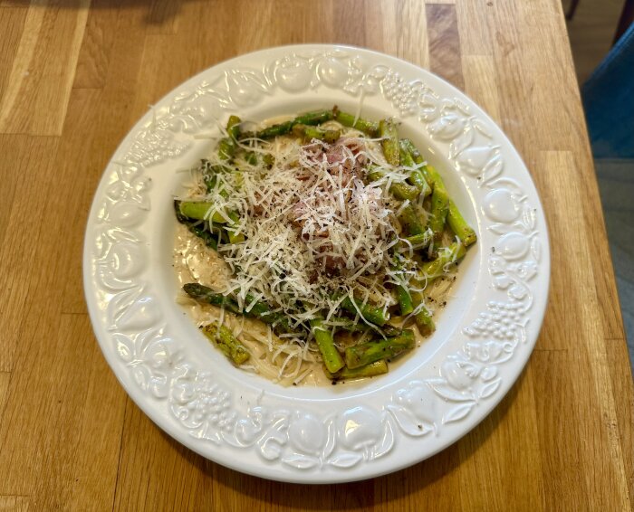 En tallrik pasta med sparris och riven parmesan på ett träbord.