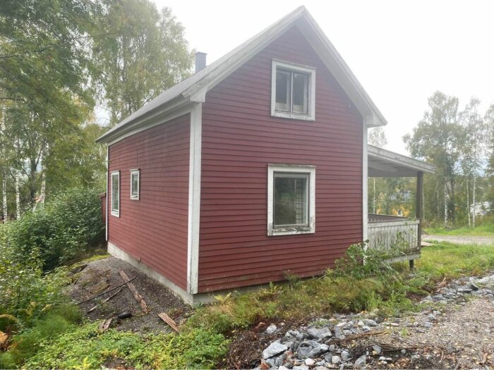 Rött trähus med två våningar, vita fönster, veranda, omgivet av grönska och grusväg.