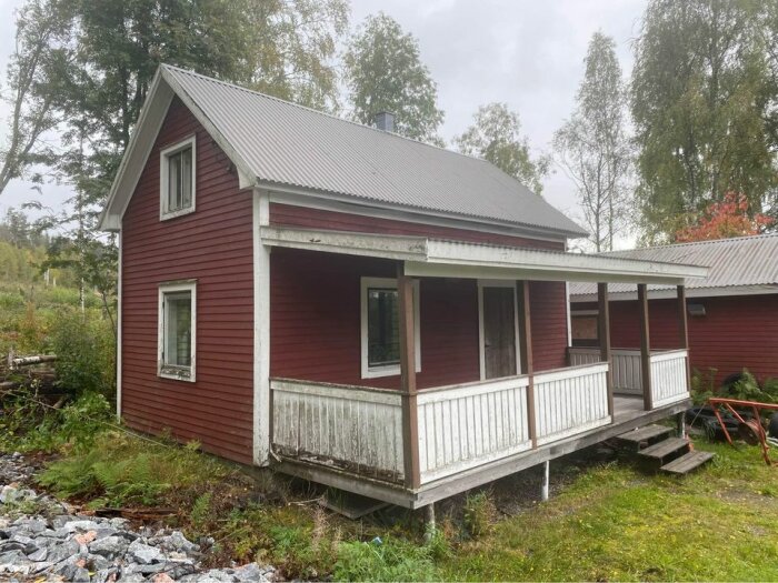 Rött hus med veranda, grått tak, omgivet av grönska, öde utseende, behöver underhåll.