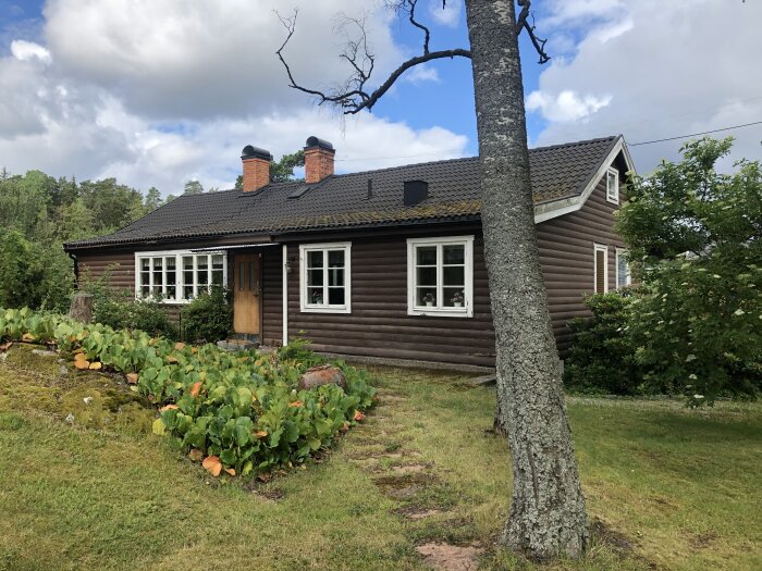 Trähus i grönska med trädgårdsland, lummig miljö, brun fasad, vita fönster, tegeltak, landlig idyll.