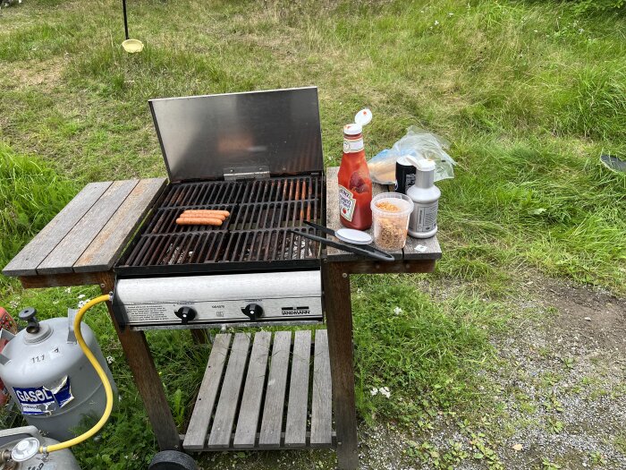 Gasolgrill utomhus med korv, ketchup, kryddor, tändvätska; gräs i bakgrunden. Grillningsscenario i trädgården.