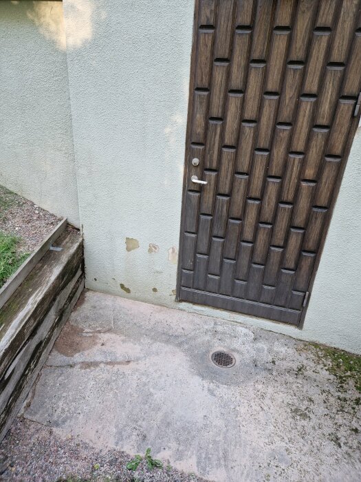 En brun dörr i trä med mönster, slitna betongtrappor, vägg med flagande färg, avloppsränna framför trappan.