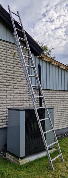 En stege lutad mot ett hus bredvid en värmepump, dagtid, grått väder, grön gräsmatta, fönster och ventilationsgaller.