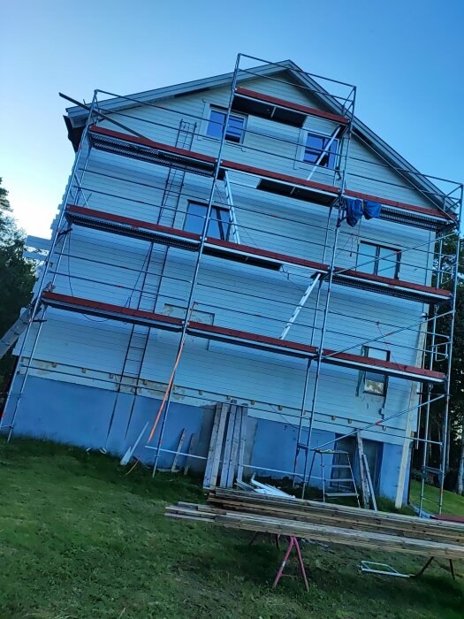 Hus under renovering med ställningar, byggmaterial på marken, delvis målat, i dagsljus.