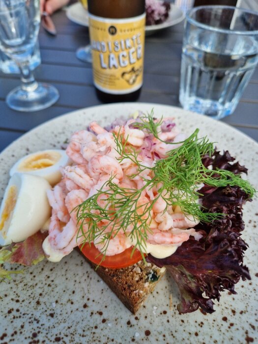 Räkmacka med ägg, dill, majonnäs, och tomat; öl och vattenglas i bakgrunden. Uteservering.