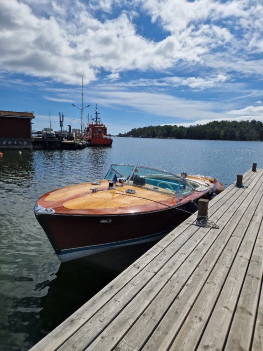 Träbåt förtöjd vid brygga, räddningsbåt i bakgrunden, blå himmel med moln, lugnt vatten.