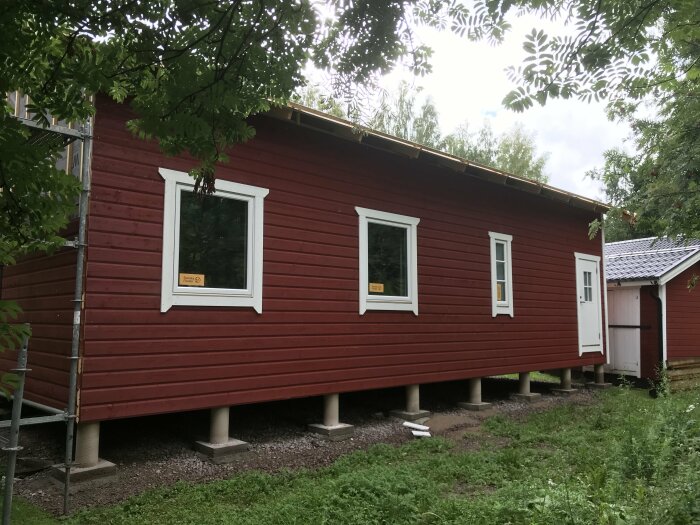 Rött trähus på pelare, vita fönster och dörr, grönska, lantligt, skyltar på fasaden.