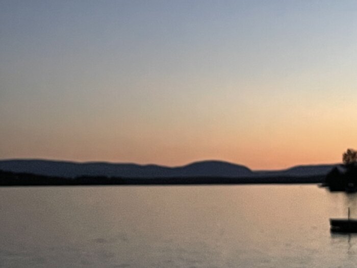 Dimmig solnedgång över sjö, silhuett av berg i bakgrunden, en båt flyter nära kusten.