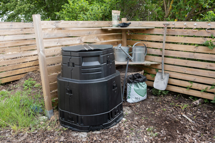 Kompostbehållare, trädgårdsverktyg, vattenkanna, jordsäck, trästaket, grönska, organisk avfallshantering, utomhus.