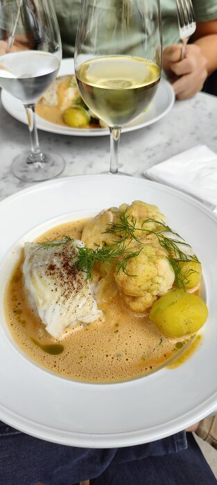 Fiskrätt med sås, blomkål och potatis, två glas vin, ätande person i bakgrunden, elegant presentation, middagsmiljö.