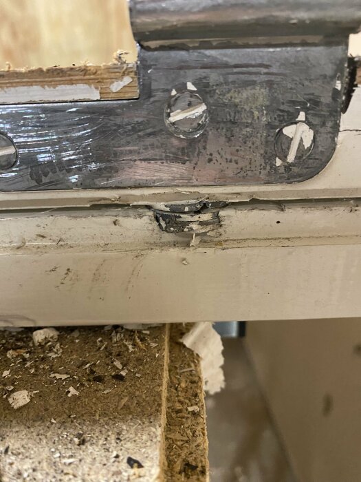 Slitet dörrstängningsbeslag på en dörrkarm, med skador och nedfallna partiklar samlade på golvet.