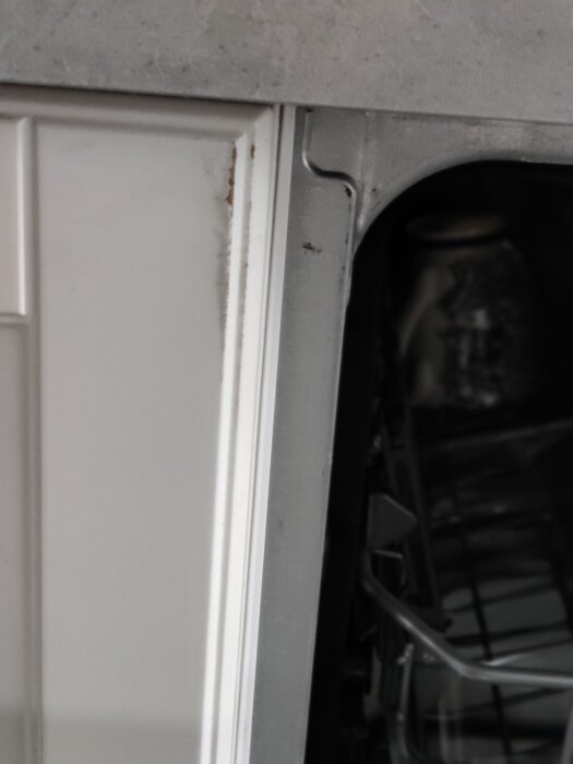 En vit dörrkant mot en grå diskmaskin, synlig förslitning och smuts, delvis öppen diskmaskinsdörr.