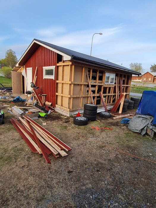 Renovering av rött trähus; stomme synlig, byggmaterial och verktyg utspridda, pågående arbete.