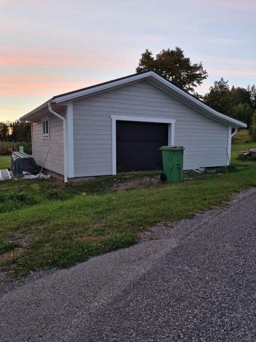 Enkel grå husfasad med garage, grönt soptunna, grusväg, kvällshimmel i bakgrunden.