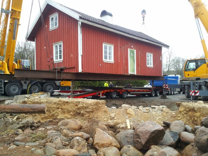 Rött trähus lyfts av kran på lastbil, flyttning av byggnad, arbetsmän och grävmaskiner på byggarbetsplats.