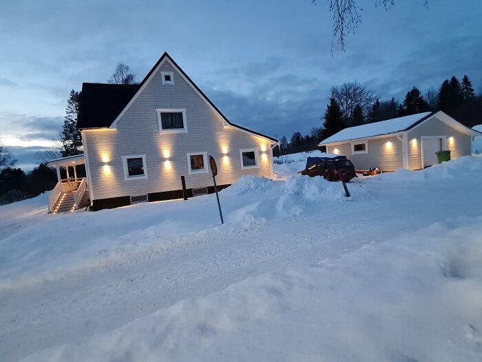 Ett hus och garage i snölandskap vid skymningen, med utomhusbelysning och parkerad bil.