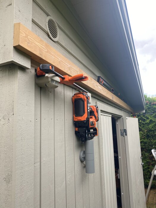 Trådlös spikpistol upphängd på en vägg under träregel vid en byggnad, nära ett ventilationsgaller.