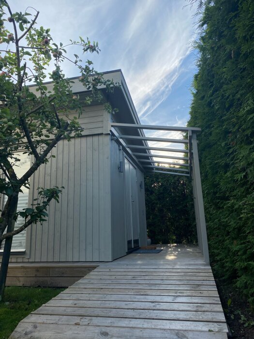 Trädäck leder till modernt hus med veranda och pergola, äppelträd och barrväxter, molnstrimmig himmel.