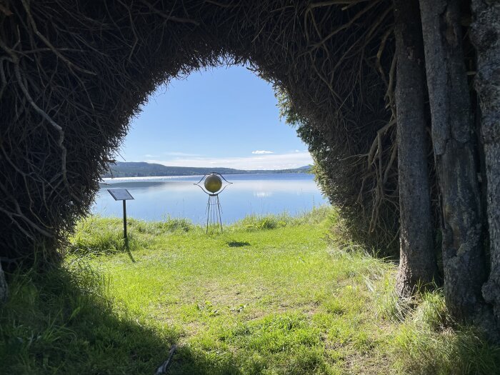 Ett naturligt valv av grenar ramar in en sjövy med en konstig skulptur och informativ skylt.