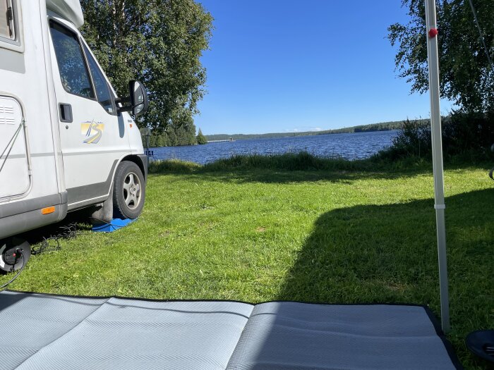 Husbil vid sjö, grönt gräs, soligt, campingliv, semester, utomhus, natur, avkoppling, blå himmel, sommardag.