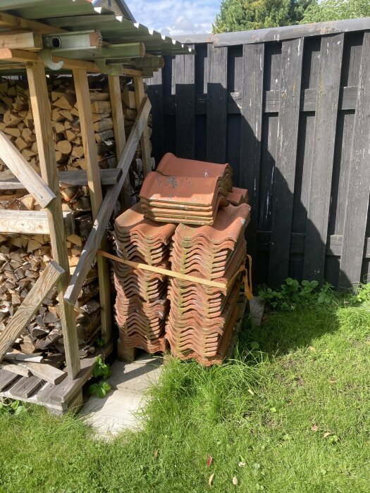 Stapelade taktegel vid vedskjul, grönt gräs, trästaket under solig himmel.