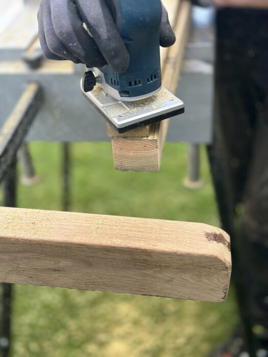 Hand med skyddshandske använder slipmaskin på träplanka; slipdamm runt omkring.