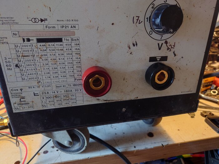 Äldre, smutsig svets omformare med justerbara reglage, tekniska specifikationer, och säkerhetsdekal på verkstadsbänk.