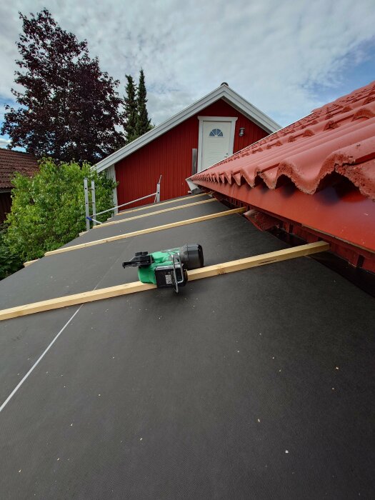 Taksäkerhetsutrustning, cirkelsåg, röd stuga, träd, molnig himmel, byggnadsarbete på tak.