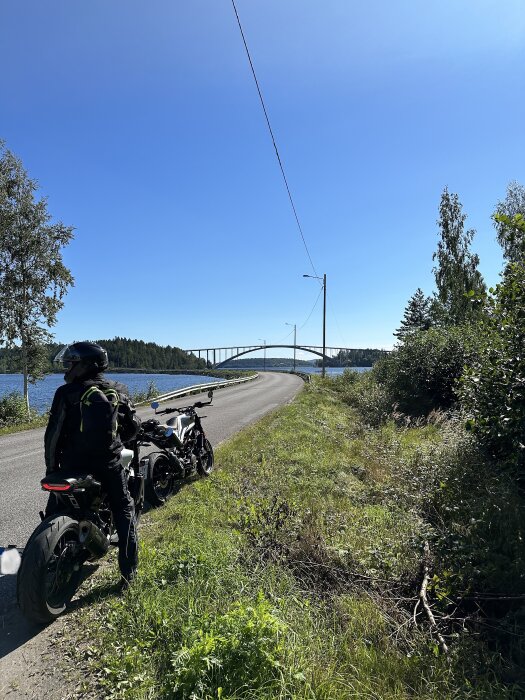 Motorcyklist och motorcykel vid vägkanten, blå himmel, grönska, bro över vatten i bakgrunden. Sunny day, outdoor adventure, road trip.