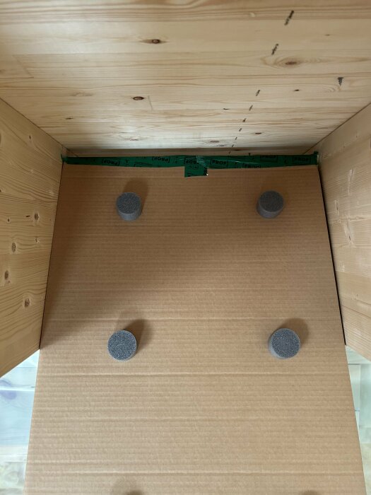 Inuti en tom låda med träkanter och möbeltassar på botten. Ljusbrun kartong, grönt tejpstycke längst upp.