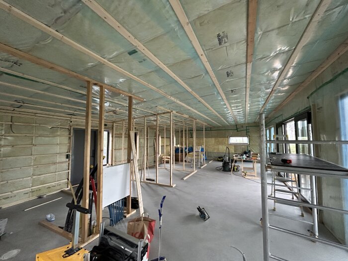 Ett pågående byggarbete inomhus med isolering, träreglar, stegar och byggnadsverktyg utspridda på golvet.