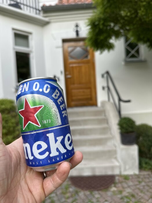 Hand håller Heineken 0.0 ölburk framför ett hus med träddörr och stentrappa.