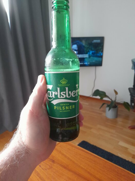 Person håller en Carlsberg Pilsner-flaska framför TV i ett vardagsrum.