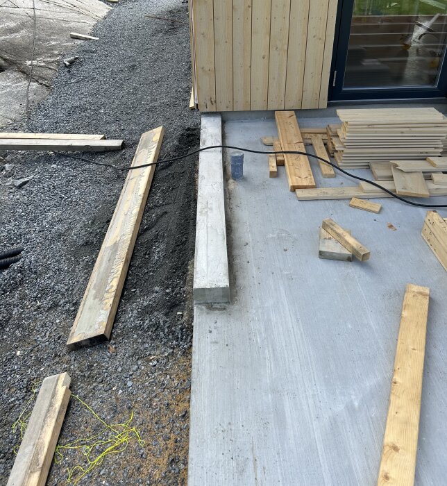 Byggarbetsplats med material och verktyg nära en nybyggd trävägg och fönster. Grus och betonggrund synlig.